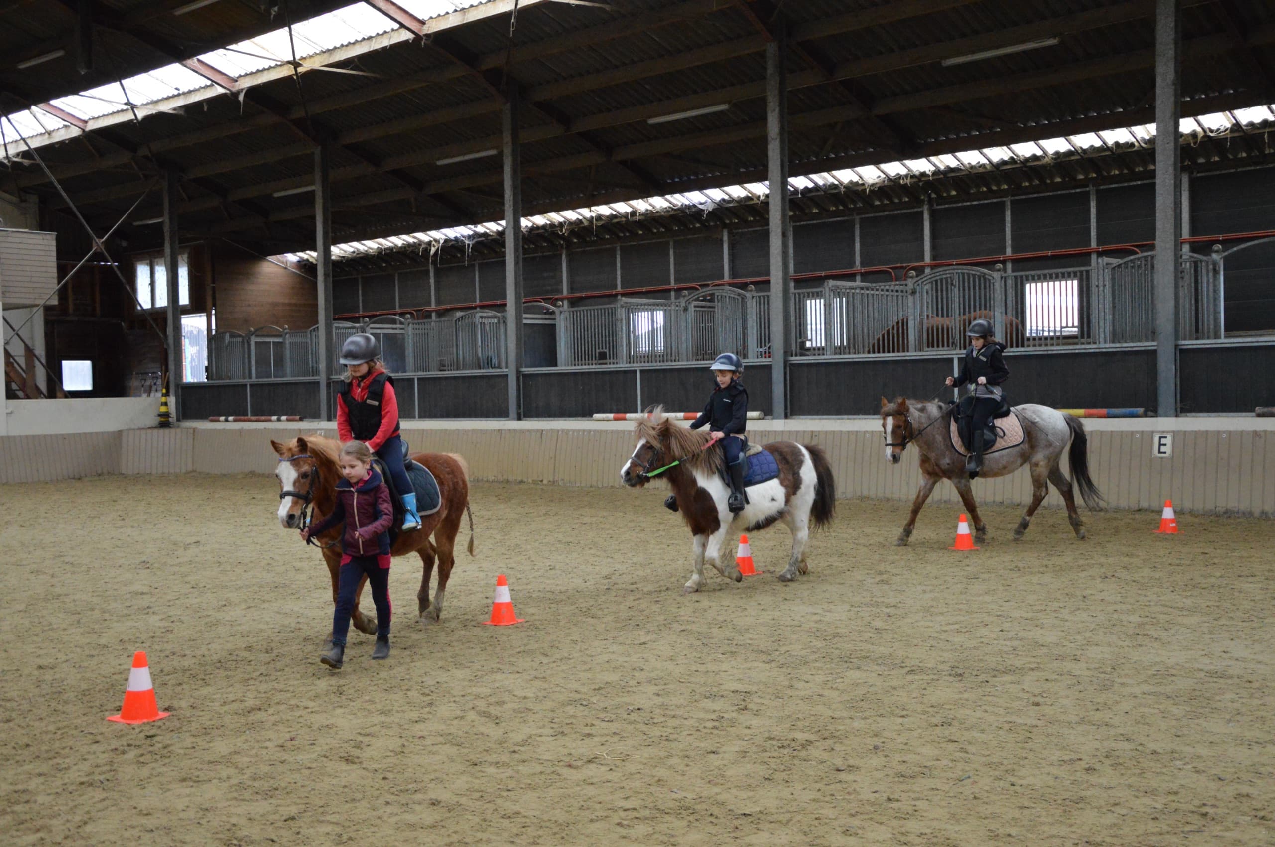 Rijles in de overdekte piste: een begeleidster leidt een pony aan de hand terwijl andere kinderen rijden